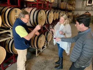 A winemaker draws wine from a barrel with a wine thief for two guests in a barrel room
