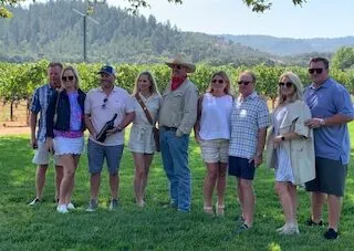 A large group of friends posing together on a winery lawn with vineyards and hills behind them