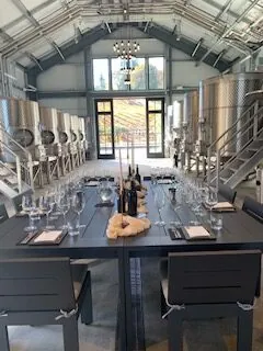 A long tasting table set with glasses and bottles inside a winery, with steel tanks along both walls